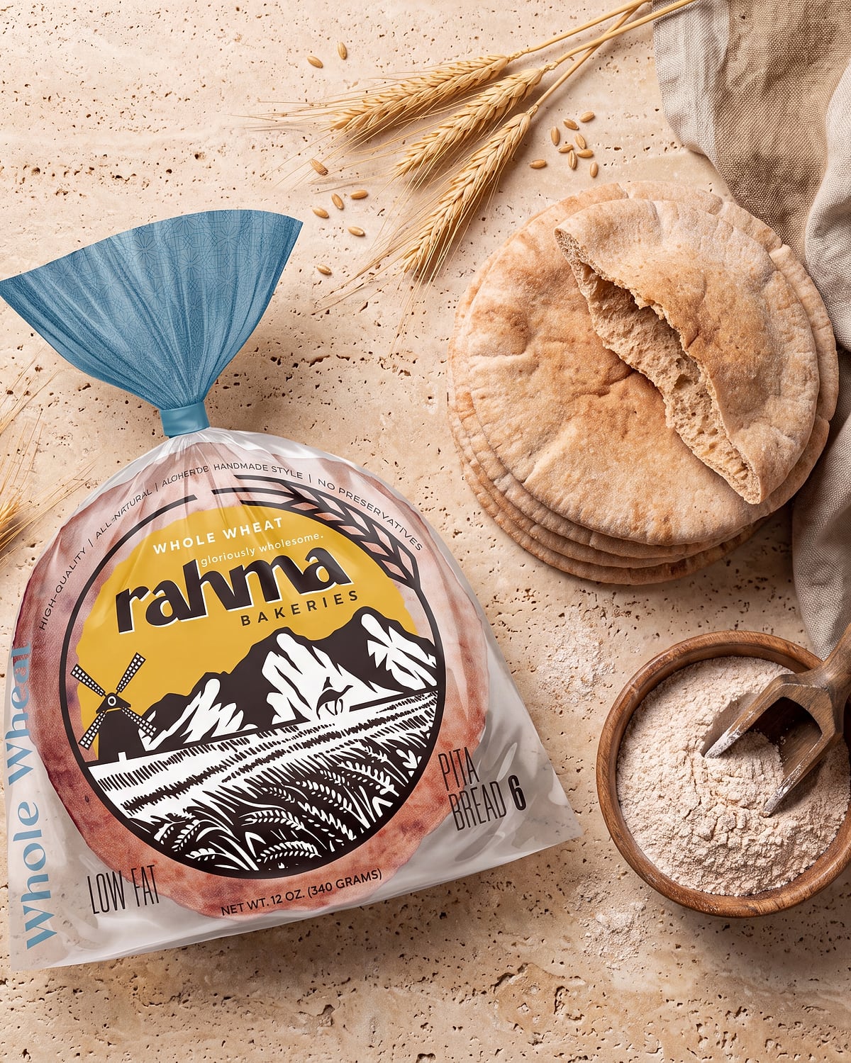 A bag of Rahma Whole Wheat pita on warm travertine, beside a stack of fresh pitas, a bowl of flour, and wheat stalks.